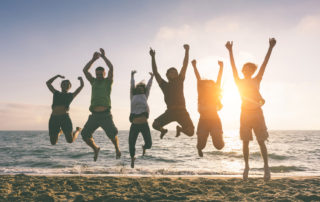 Group of 6 people at the beach jumping mid air with the sunset and ocean behind them